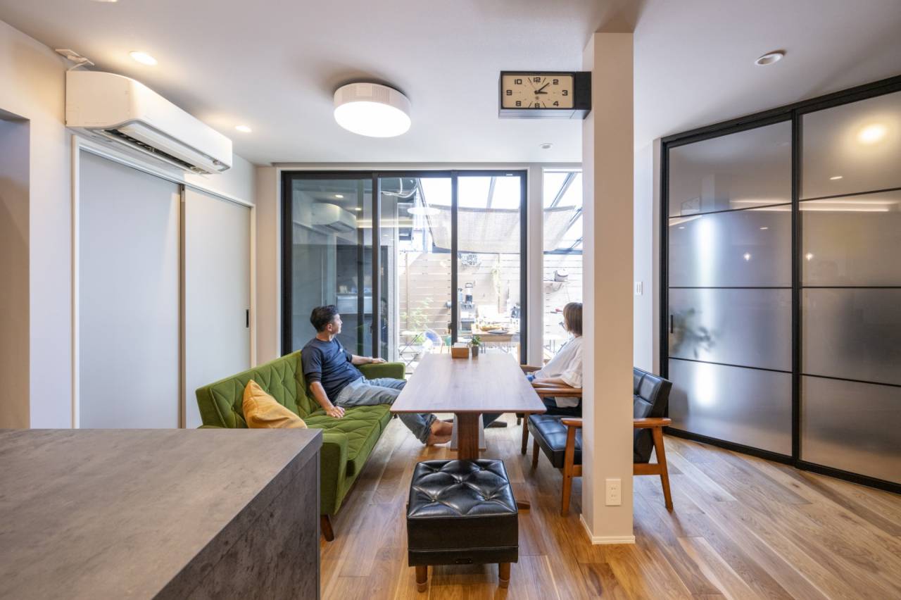 リビングとフラットに繋がるタイルテラス（アウトドアリビング）のある平屋。防犯性能を高めた外構壁に囲まれたテラスで、外からの視線を気にせずくつろぐ夫婦。室内はグリーンソファと木目テーブル、奥には半透明のハンガー引戸。パッシブデザインによる自然採光。