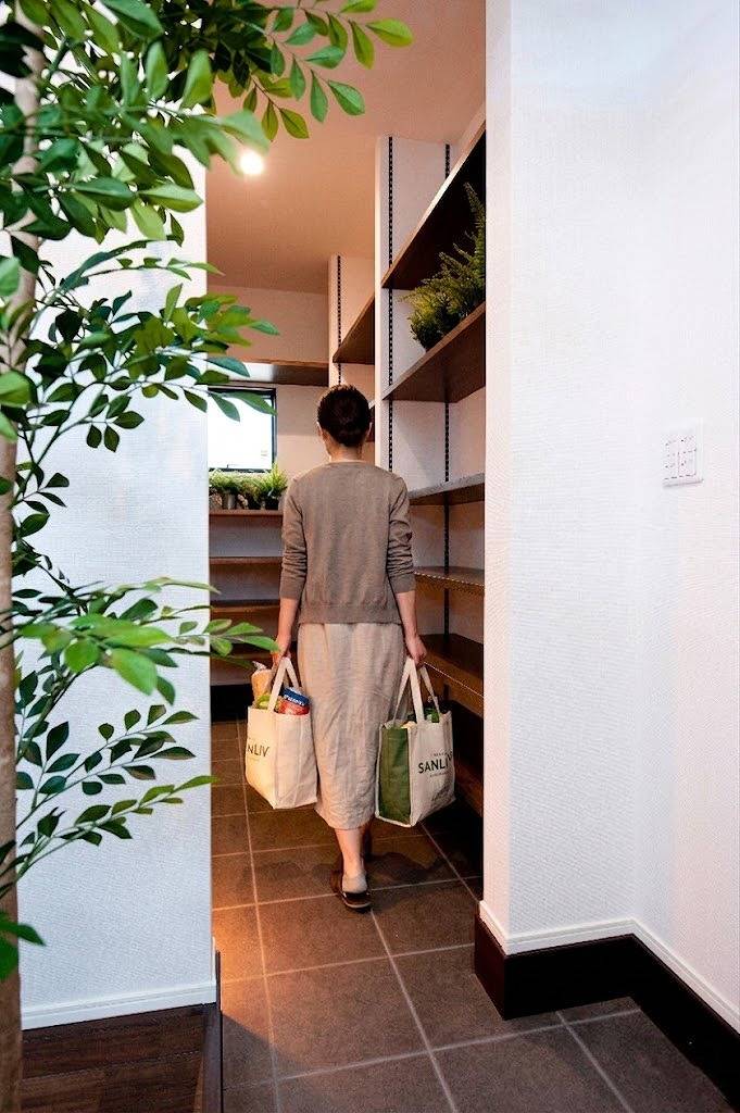 玄関からキッチンへ続く土間収納（パントリー）の様子。買い物袋を両手に持った女性が、可動棚が設置されたゆとりある通路を通るシーン。重い荷物をすぐに置ける、家事動線に配慮した住宅設計の実例